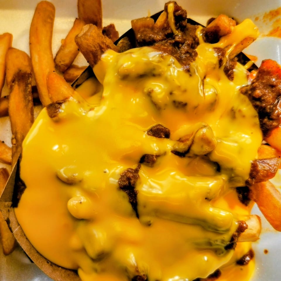 Close-up of chili cheese fries in a white takeout container, overflowing with melted cheese and chili.