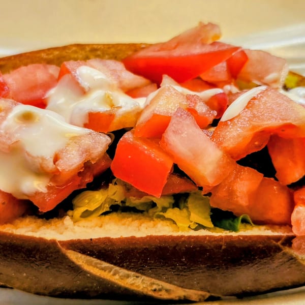 Close-up of a pretzel bun sandwich filled with lettuce, meat, diced tomatoes, and white sauce.
