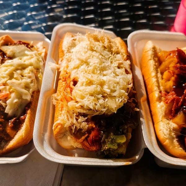 Three hot dogs with various toppings, including chili, coleslaw, sauerkraut, and bacon, in white foam containers.