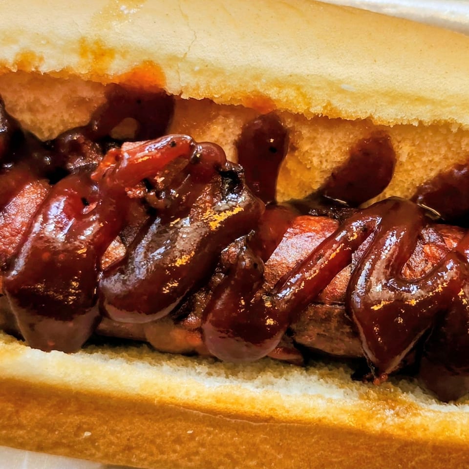 Close-up of a hot dog in a bun, generously topped with dark, glistening barbecue sauce.