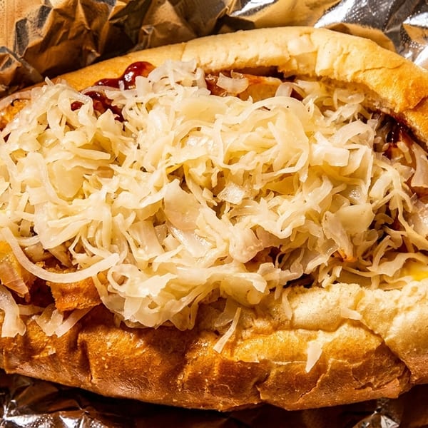 Close-up of a hot dog in a bun, topped with sauerkraut, onions, and sauce, wrapped in foil.