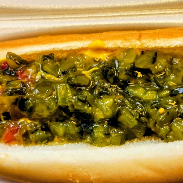 Hot dog in a bun with mustard and green relish, served in a white takeout container.