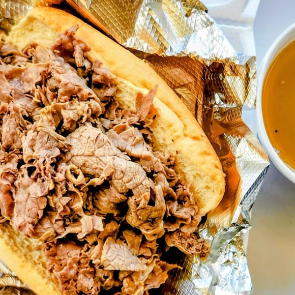 A large Italian beef sandwich in foil next to a cup of au jus.